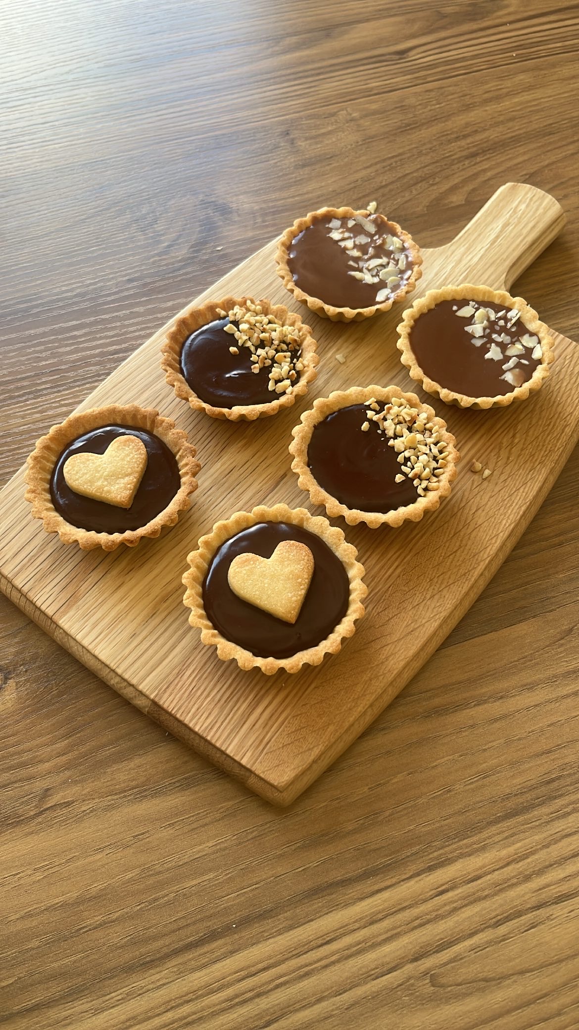 Six small chocolate tarts: two with heart-shaped cookies on top, two with almond pieces, and two with hazelnut pieces.